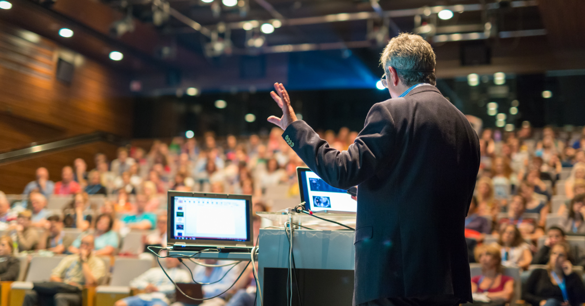 Seorang pembicara sedang menyampaikan presentasi di depan audiens dalam sebuah seminar besar yang dikelola oleh event planner.